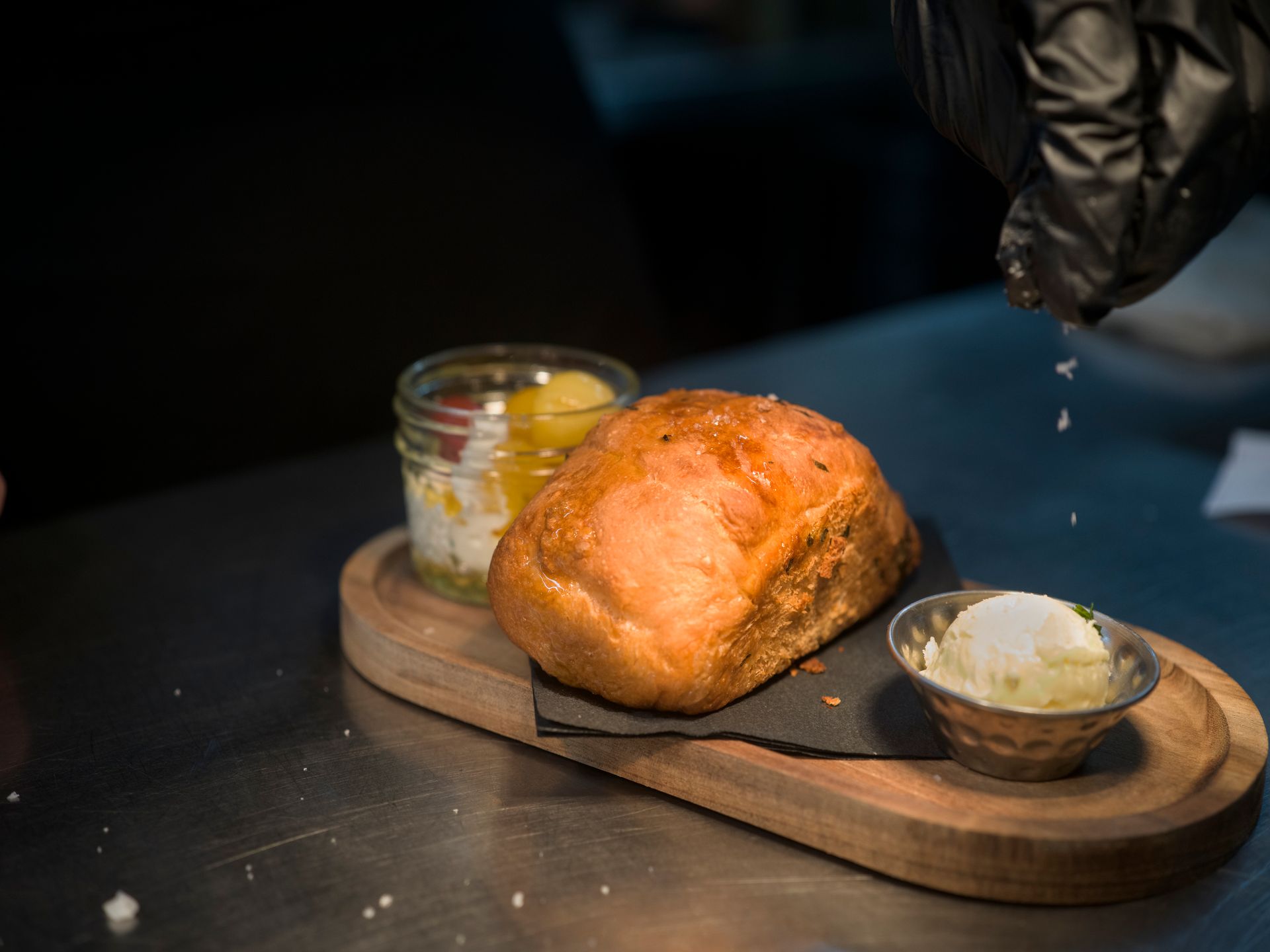 Bread loaf and two small dishes on a wooden board, with a gloved hand sprinkling salt above them.