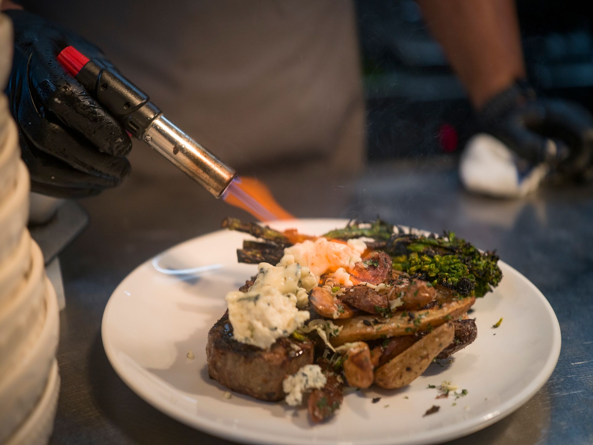 Chef torching a plated steak with mashed potatoes and greens in a kitchen