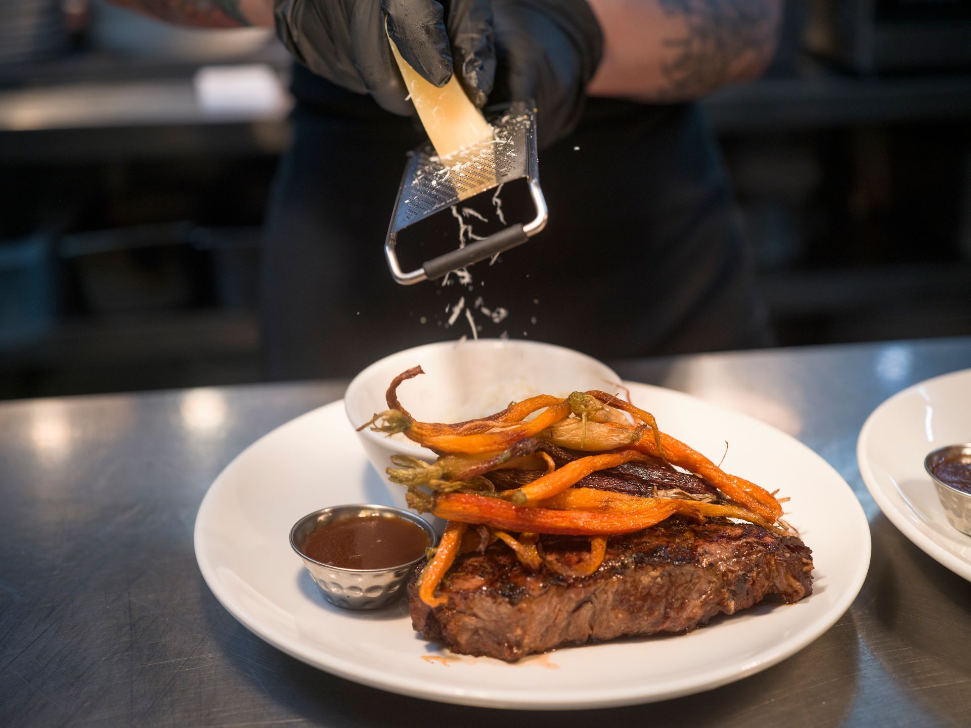 Chef grating cheese over a plated steak with carrots and sauce in a restaurant kitchen