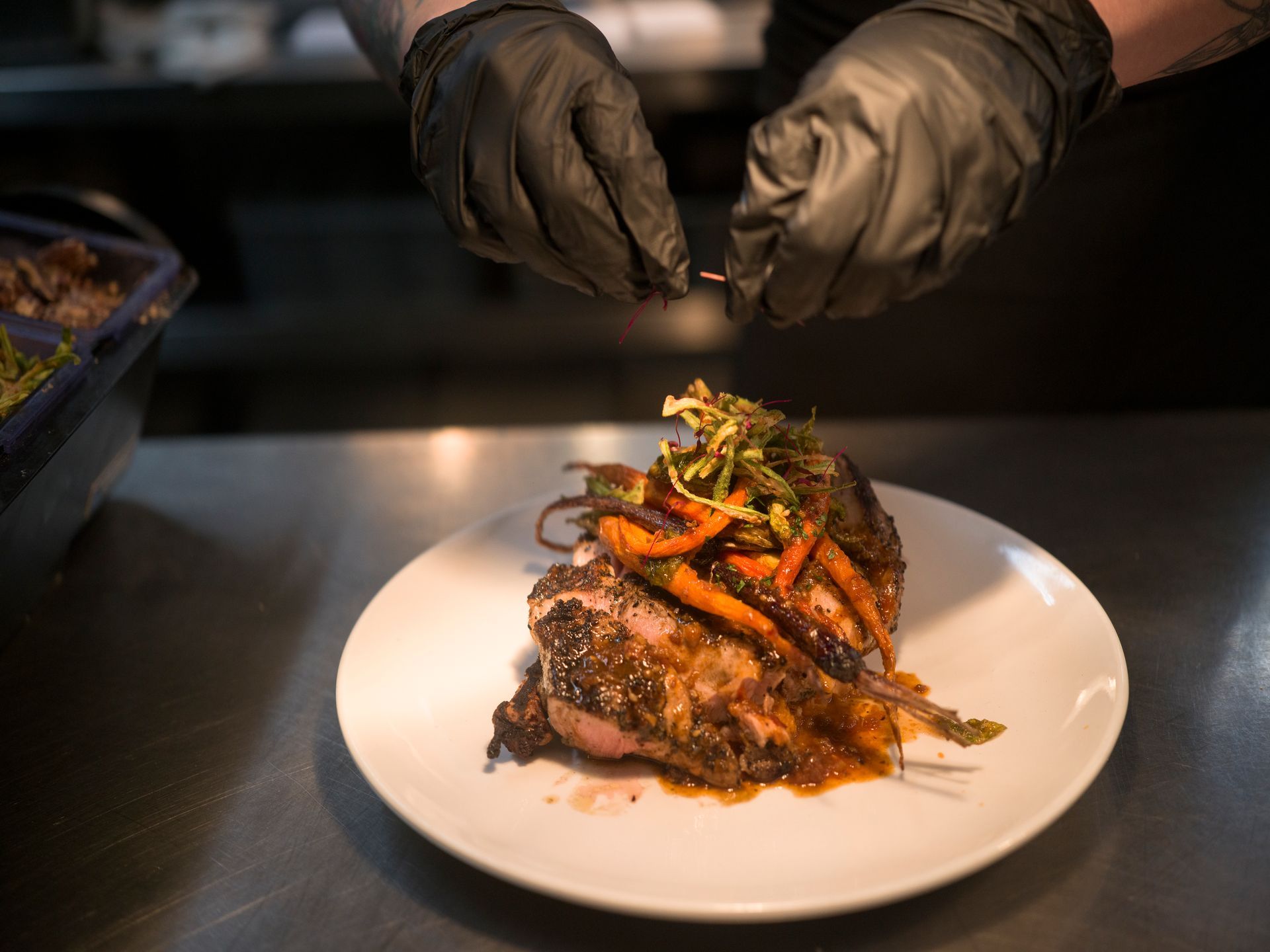 Chef’s gloved hands plating a gourmet dish on a white plate with vegetables and sauce