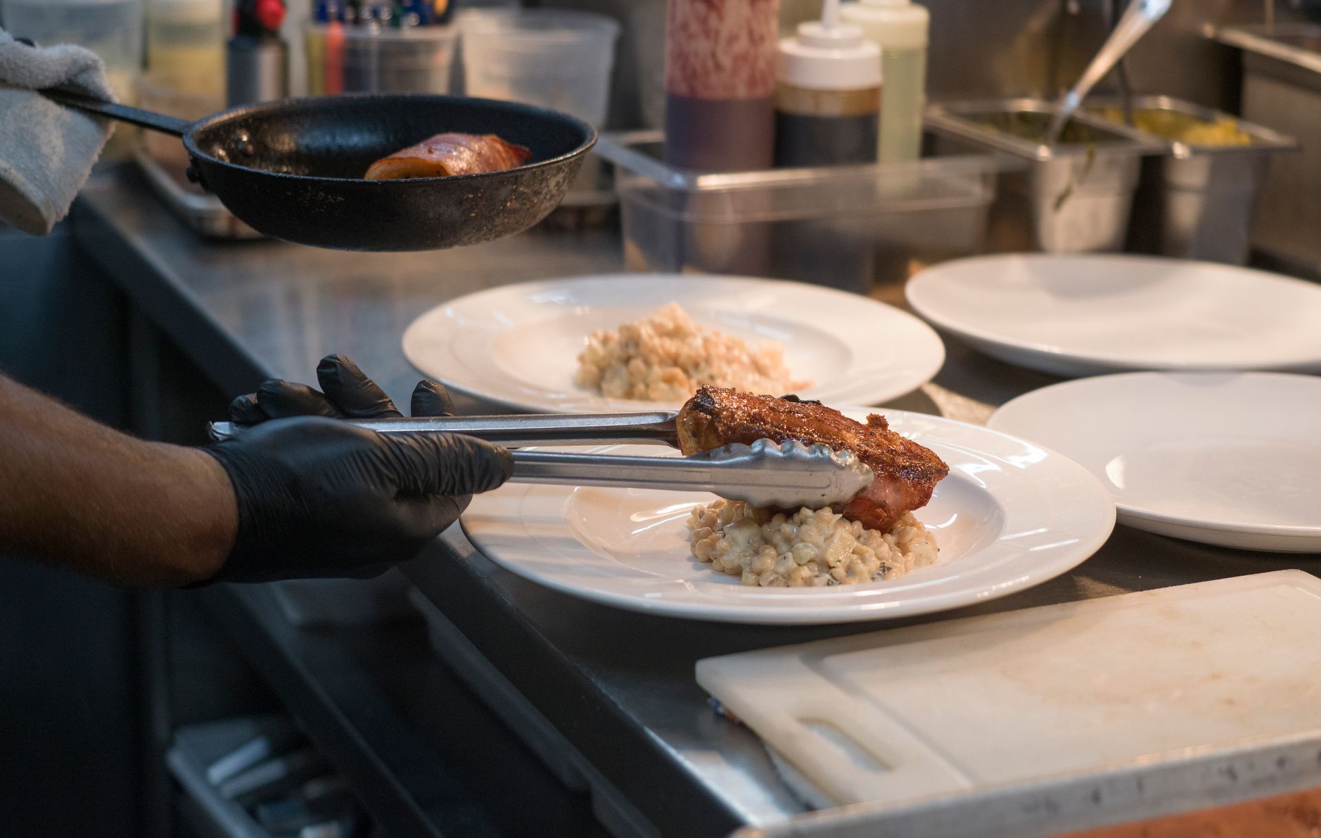 Chef plating food on white plates in a restaurant kitchen, with a frying pan and utensils nearby