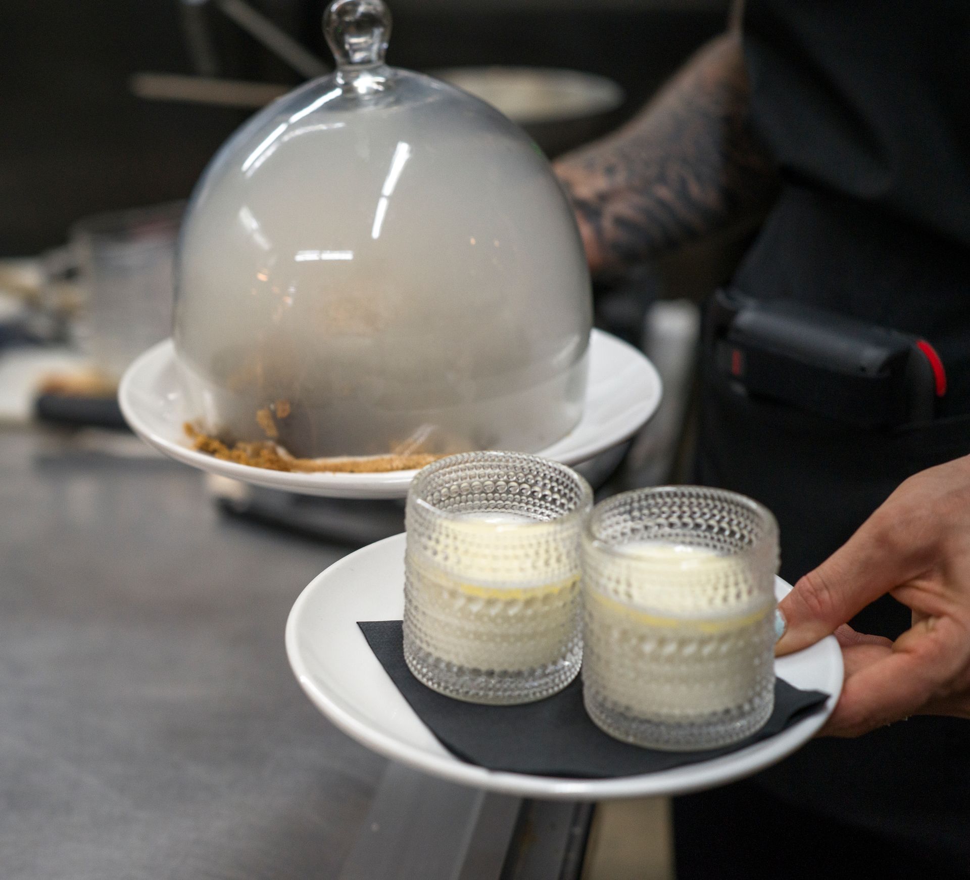 Two small glasses of creamy dessert on a white plate beside a glass cloche on a tray