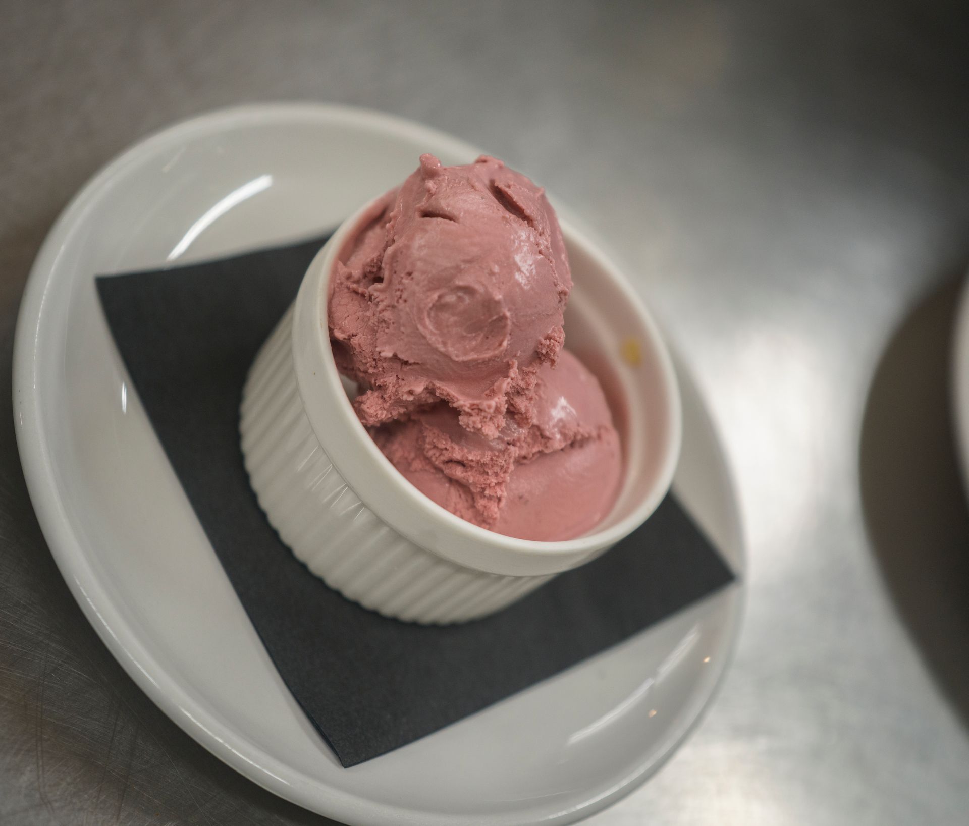 Pink ice cream in a white ramekin on a black napkin atop a white plate