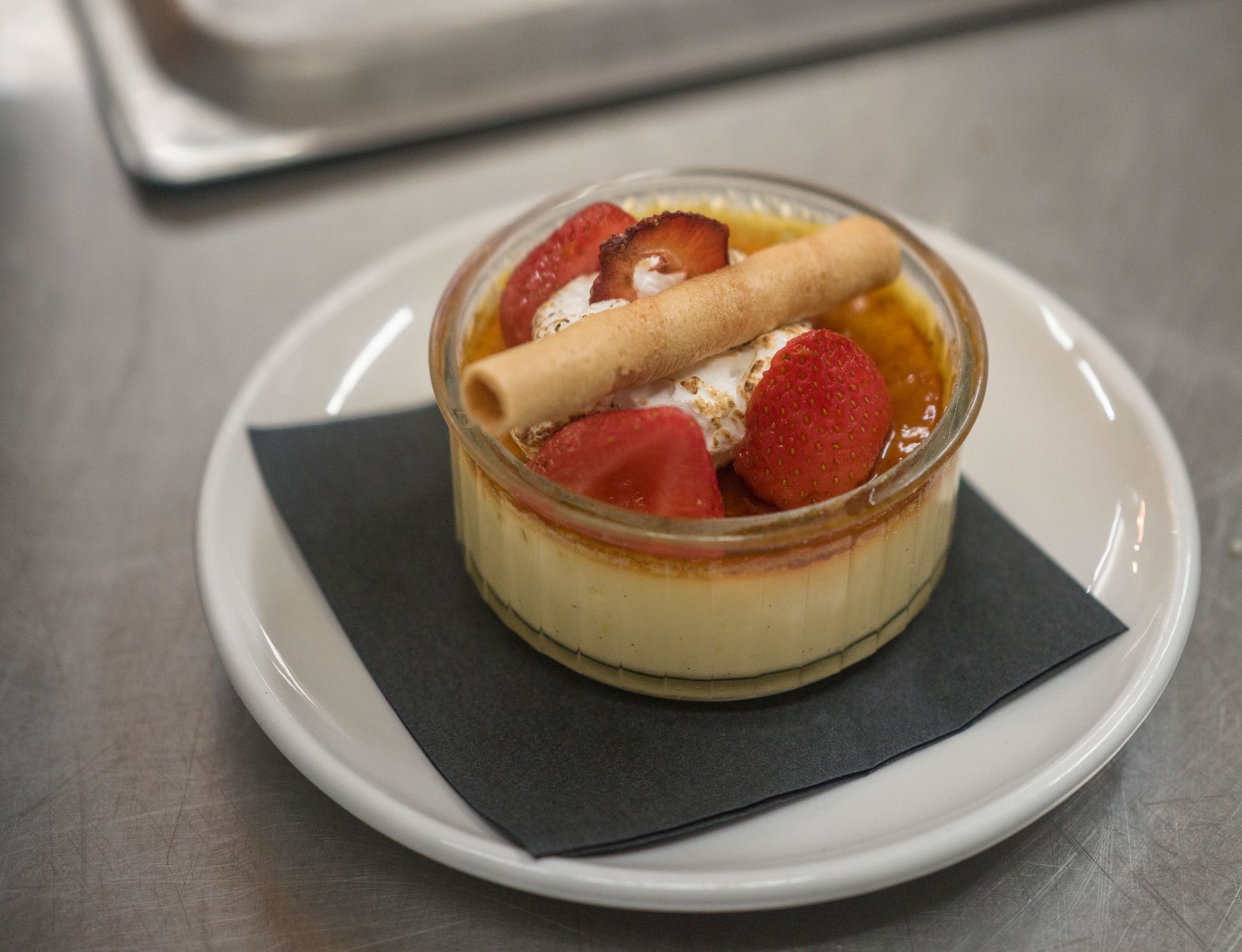 Fruit tart in a clear ramekin on a white plate with a dark napkin on a metal table