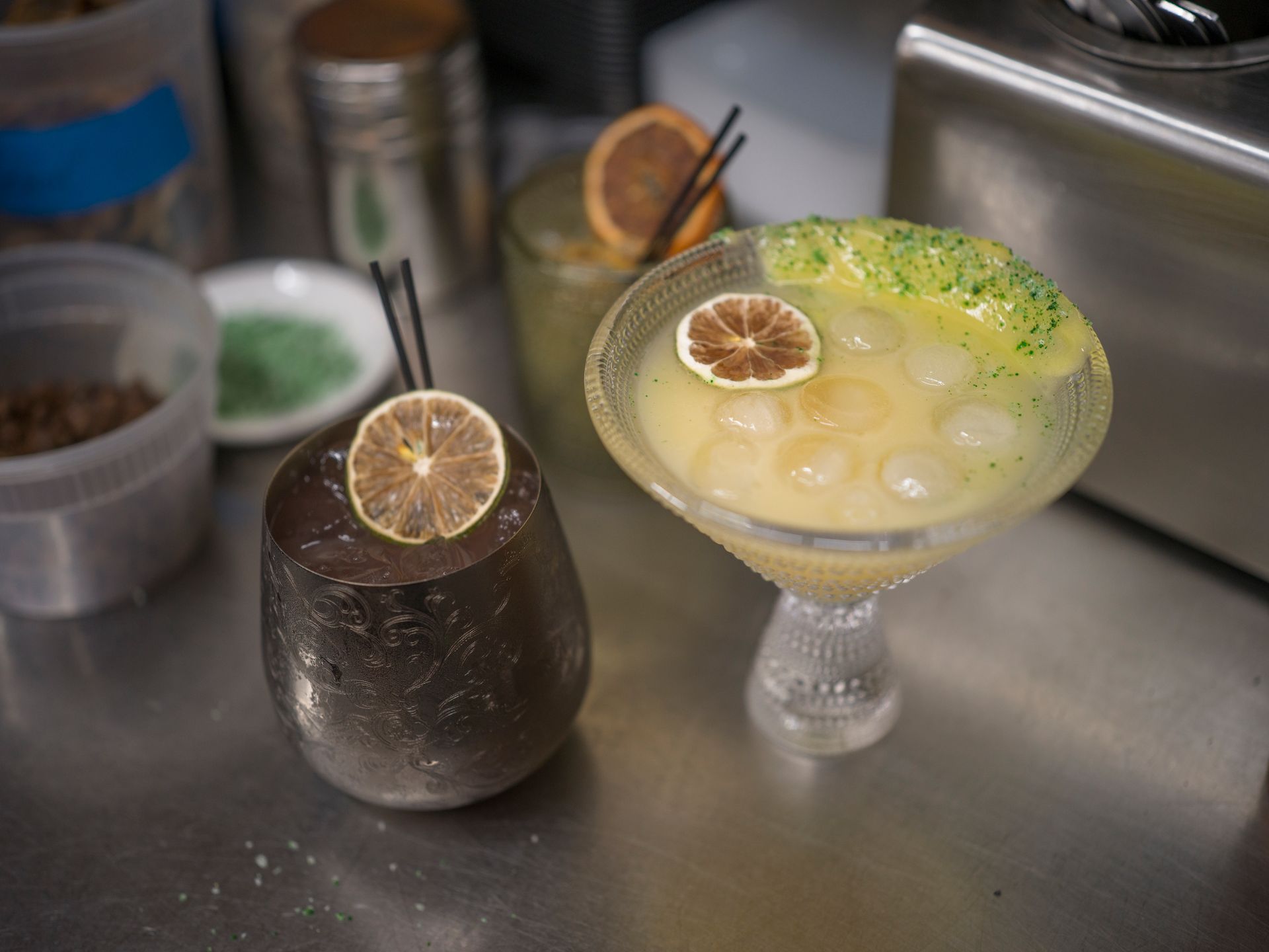 Two cocktails on a metal counter, one dark and one pale, each garnished with a dried citrus slice.