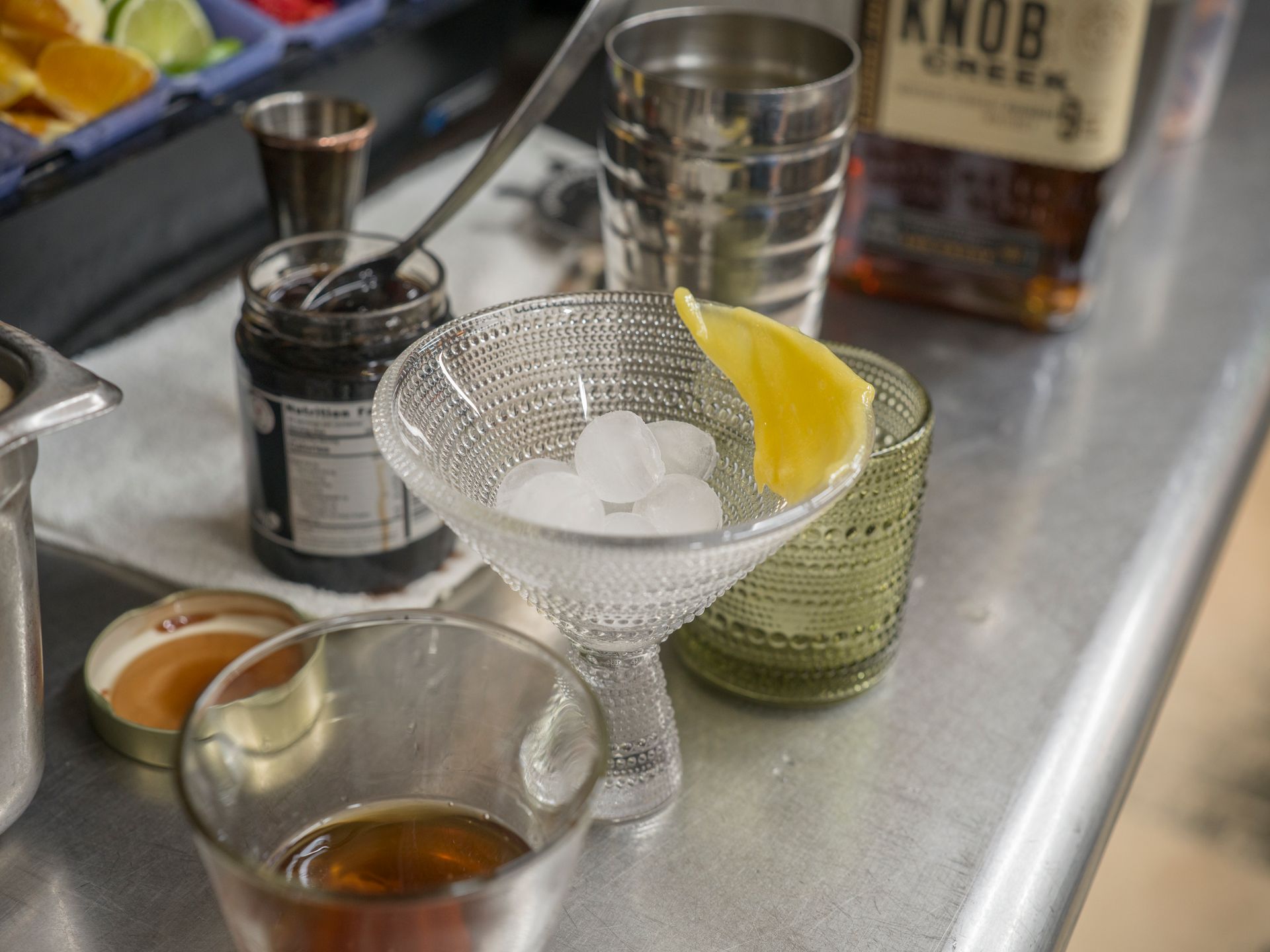 Cocktail ingredients and bar tools on a stainless steel counter, with a lemon wedge and ice in a strainer.