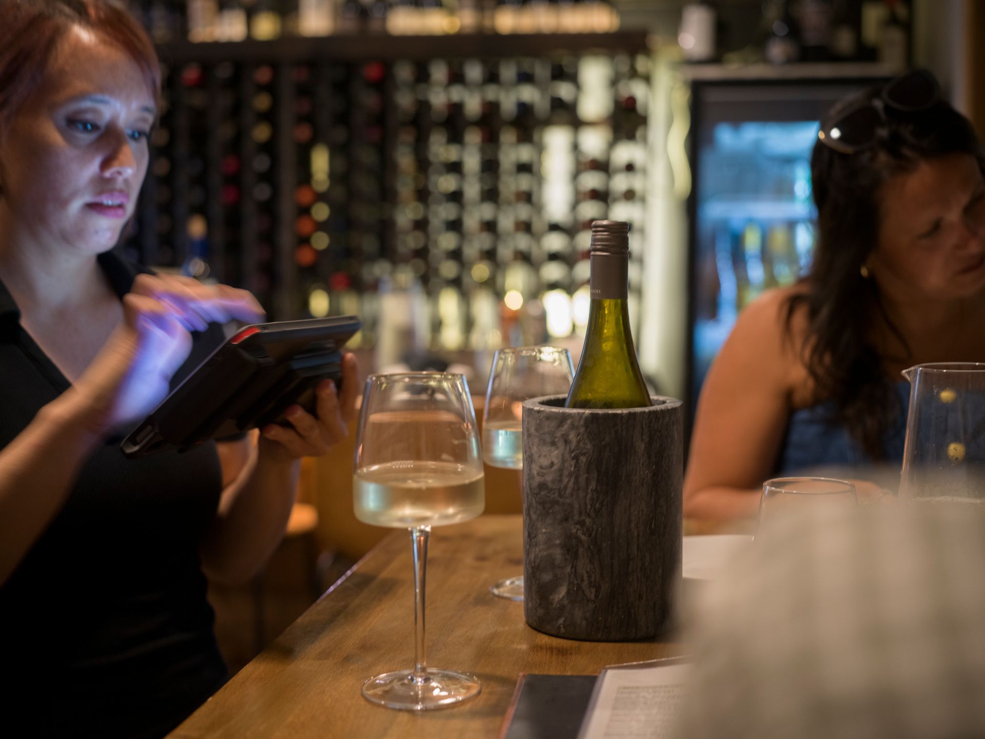Two people seated at a bar, one using a tablet beside a wine glass and bottle