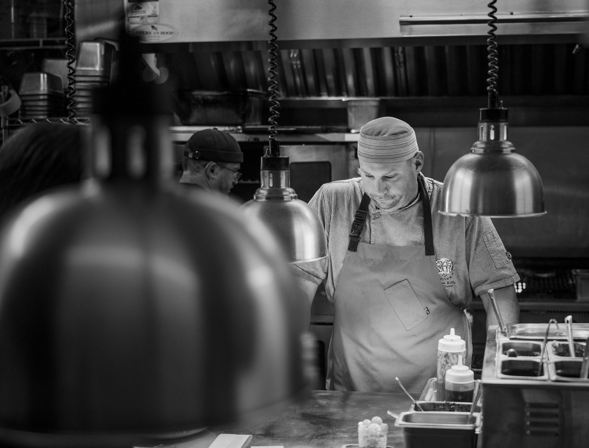 Chef in a busy kitchen, focused on food prep beneath hanging lights and steam.