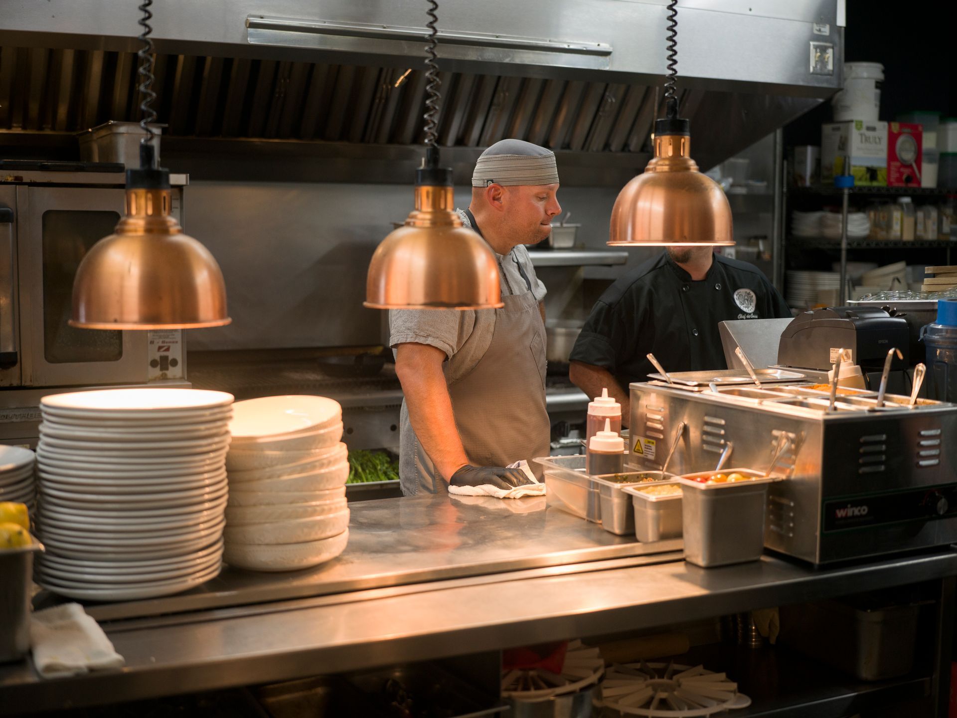 Chefs working behind a stainless-steel restaurant counter with stacks of plates and heat lamps