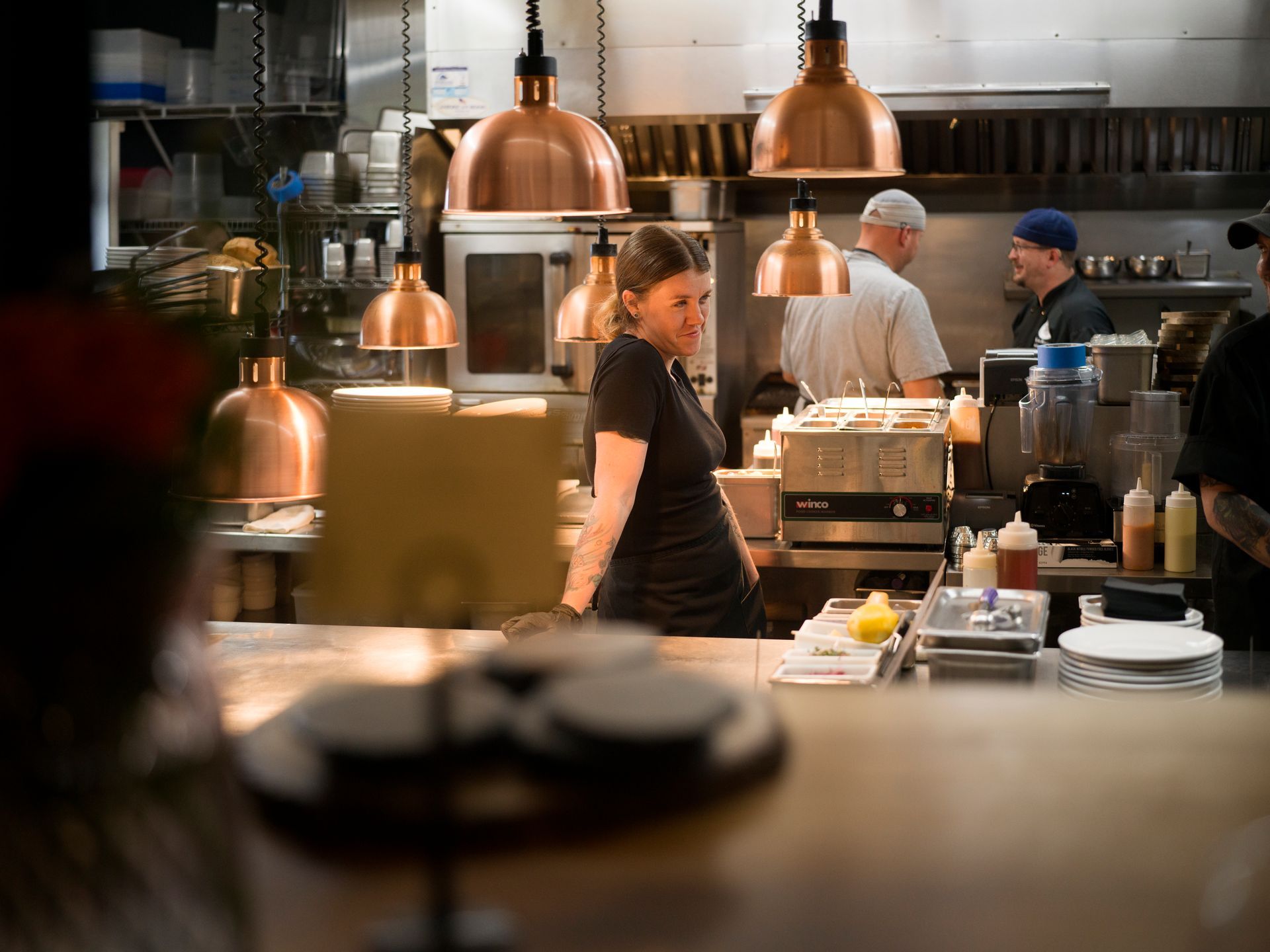 Busy restaurant kitchen with chefs working under copper pendant lights and stainless steel counters