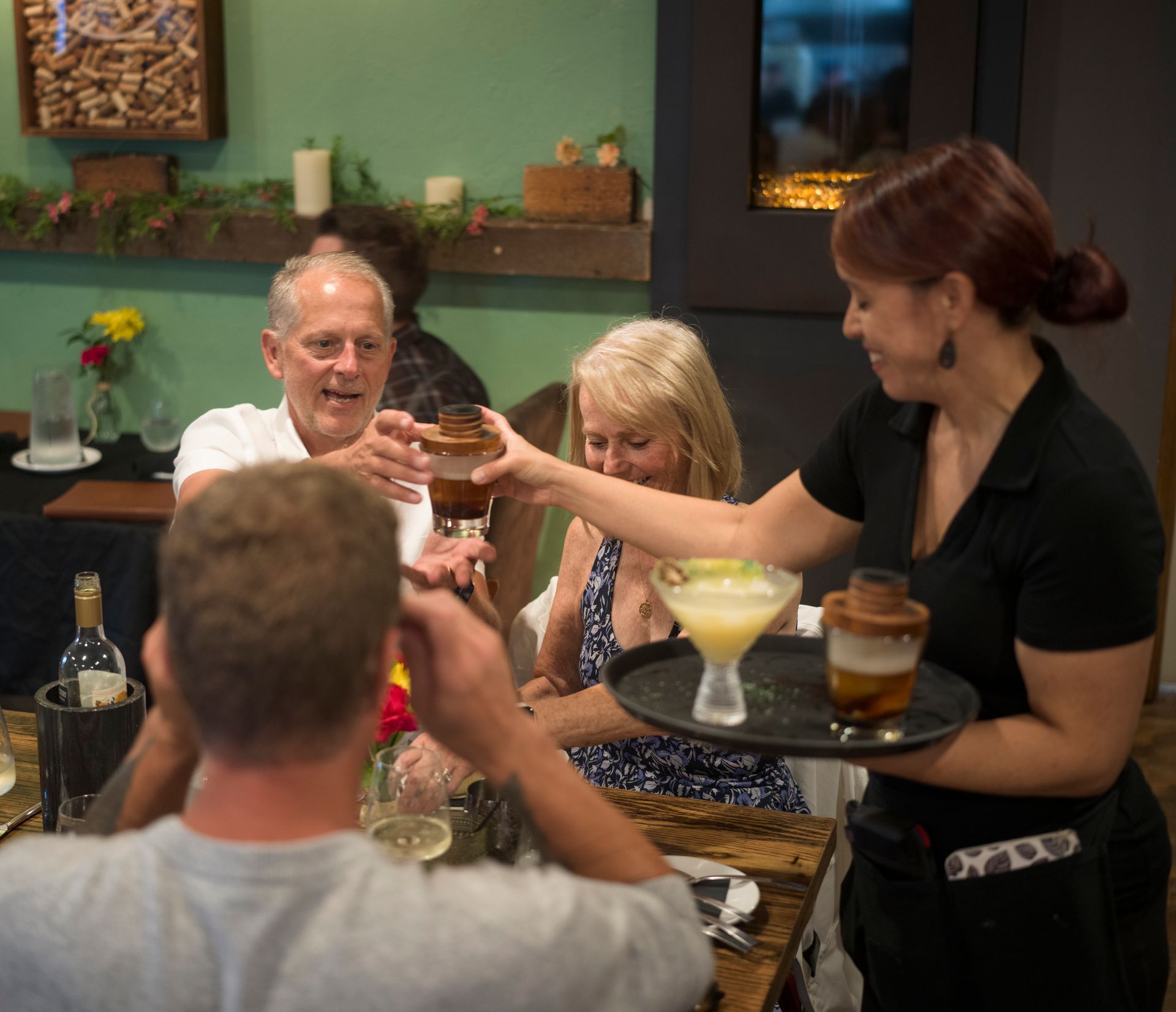 Friends toasting cocktails at a restaurant table with a server holding a tray.