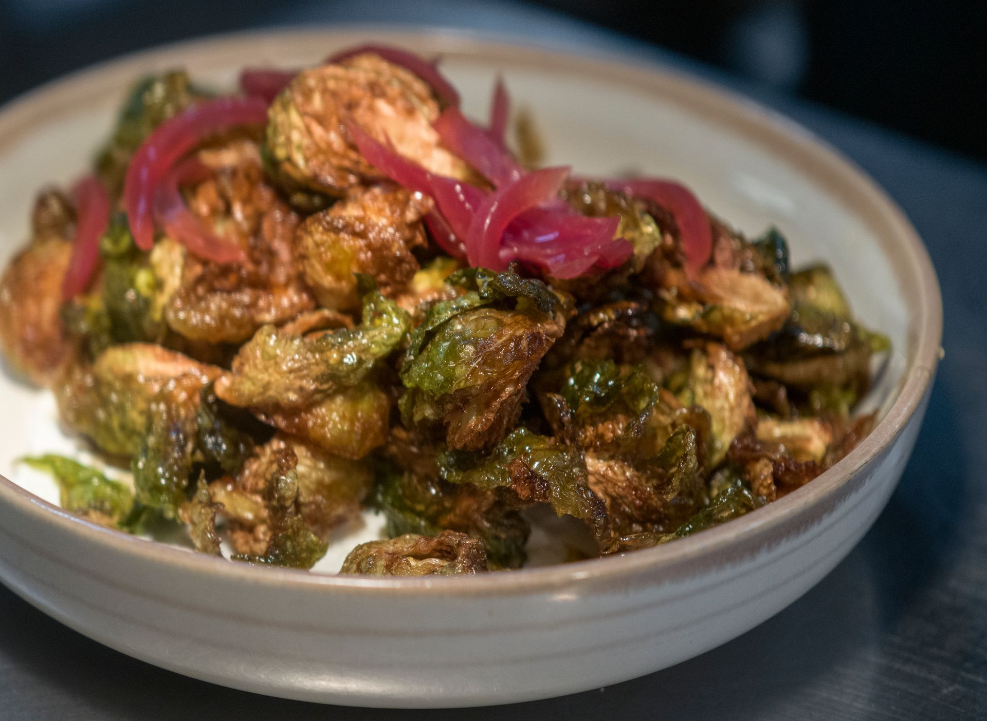 Bowl of crispy Brussels sprouts topped with pink pickled onions on a dark table