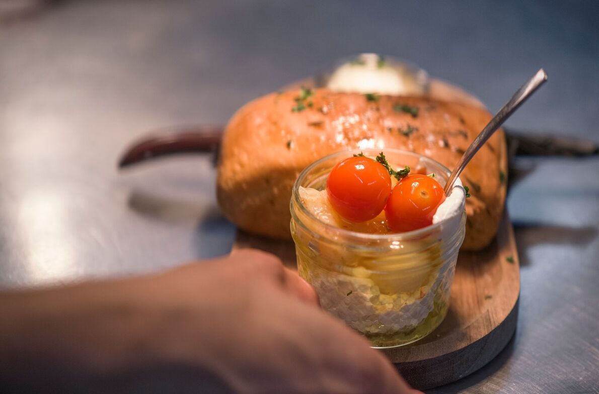 Jar of creamy dessert topped with cherry tomatoes on a wooden board, with bread in the background