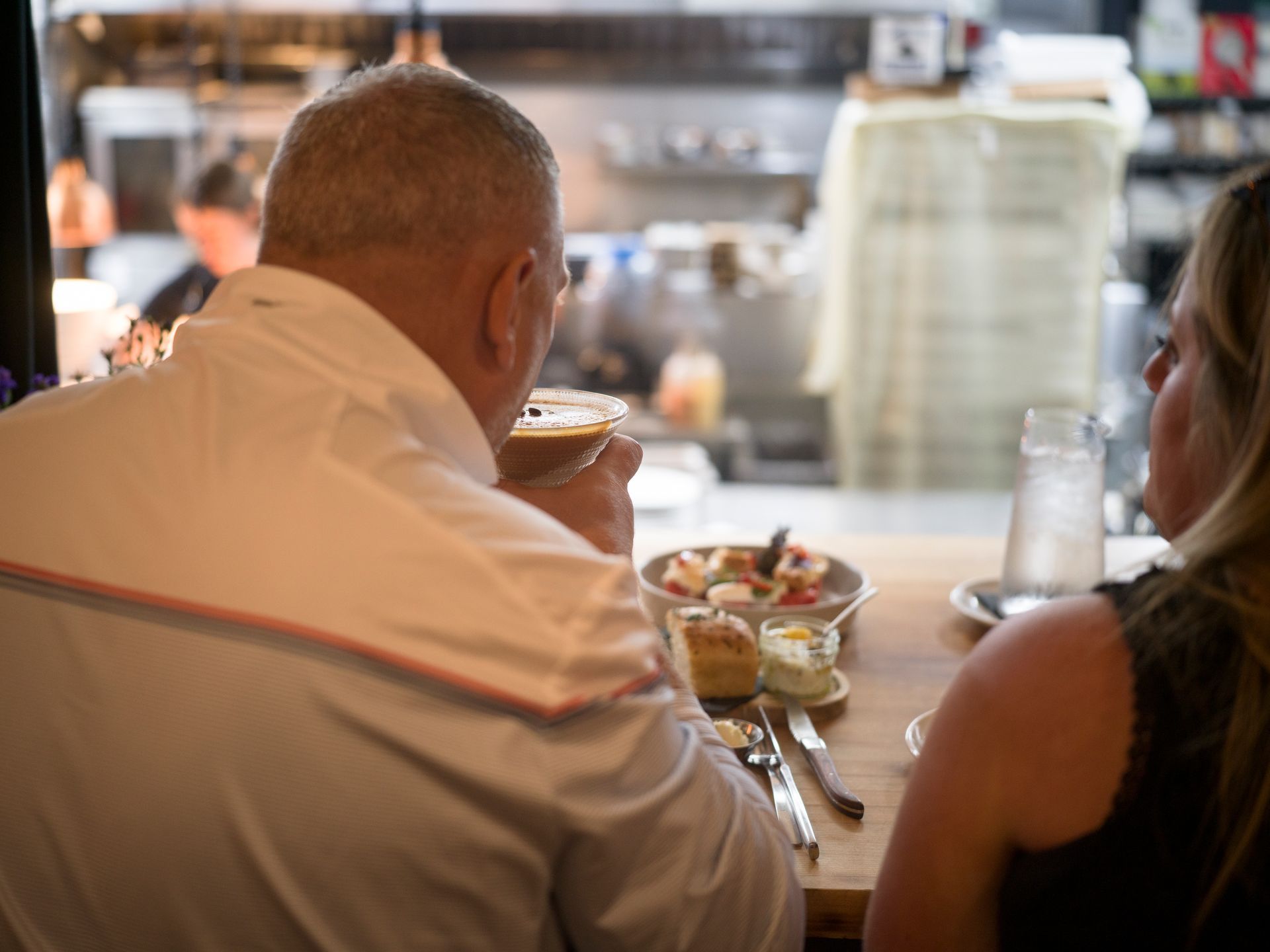 Two people seated at a diner counter, eating and chatting over drinks and plates of food.
