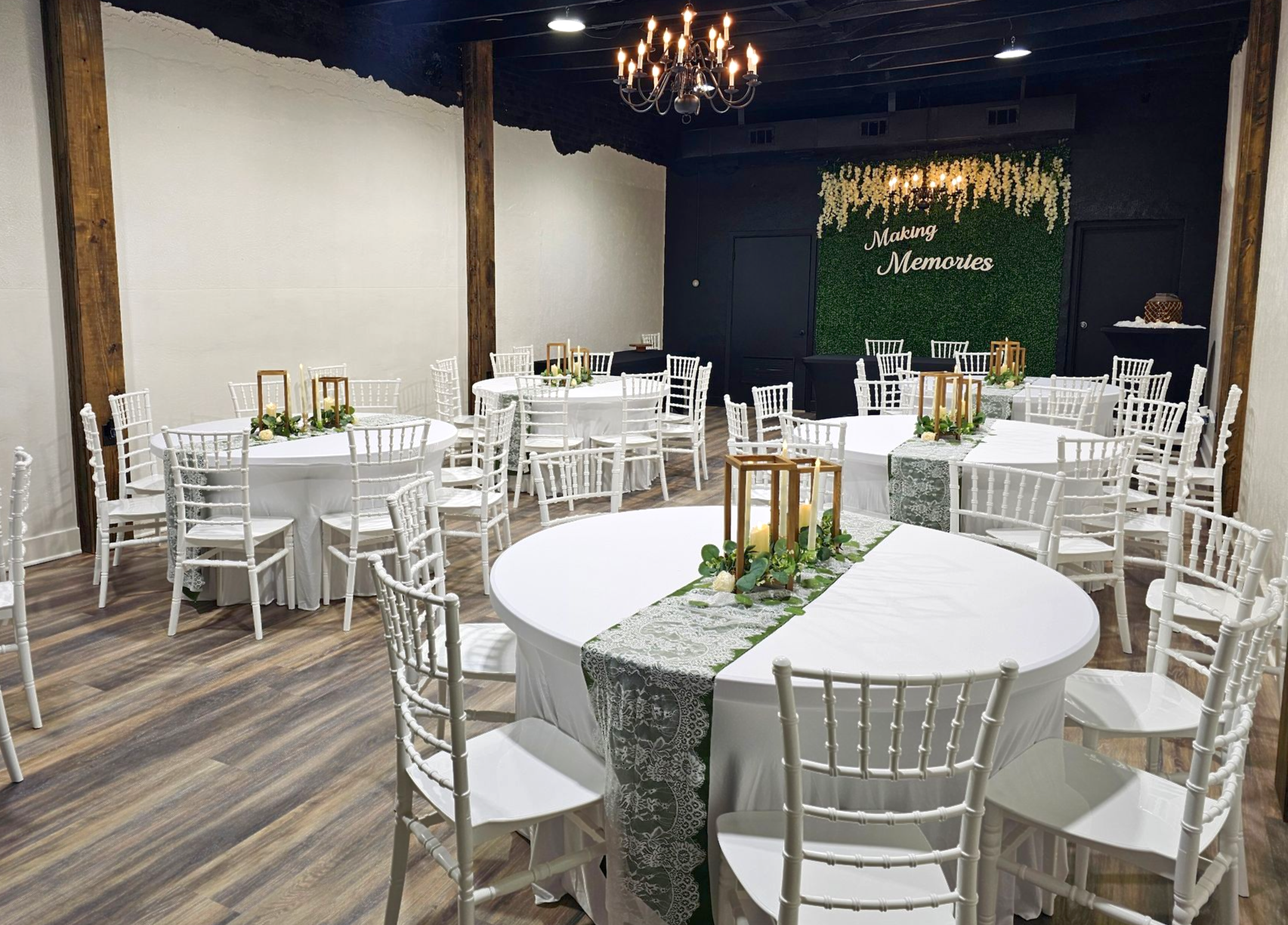 Event space featuring round tables with white tablecloths, greenery runners, and white chairs in a rustic, modern hall.