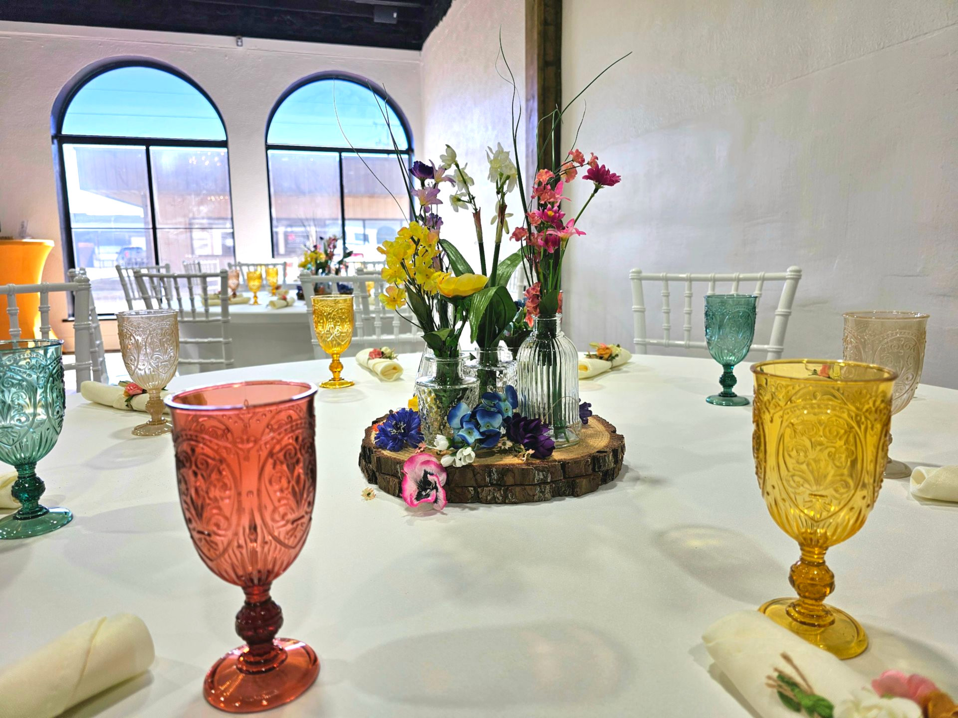 A table set for a formal event with white tablecloths, colorful glass goblets, and a central floral centerpiece on wood.