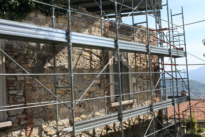 Stone wall under construction with metal scaffolding beside a tiled-roof village and distant hills