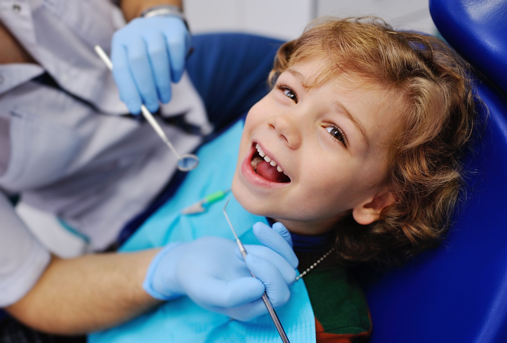 A close-up of a smiling child sitting in a blue chair with a dentist holding dental tools. A close-up of a smiling child sitting in a blue chair with a dentist holding dental tools.