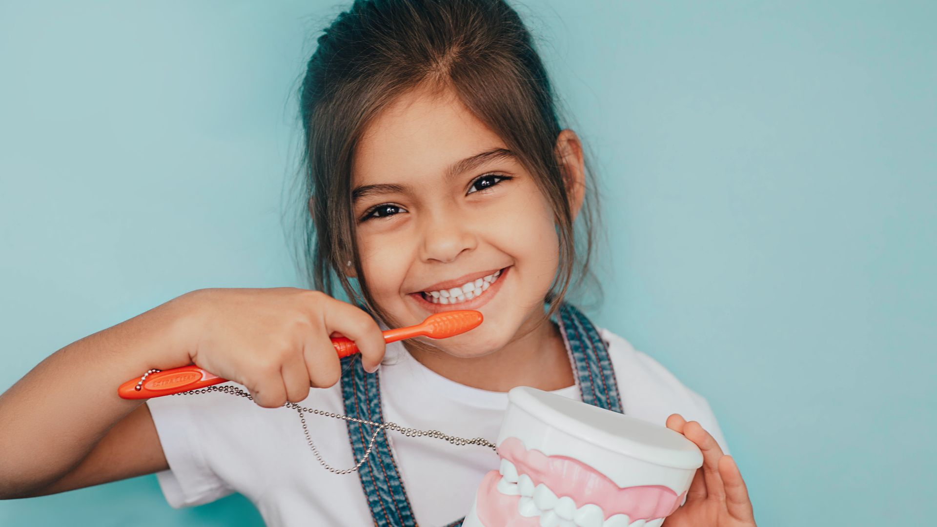 Girl brushing teeth with an orange toothbrush and holding a model set of teeth, smiling against a blue background.