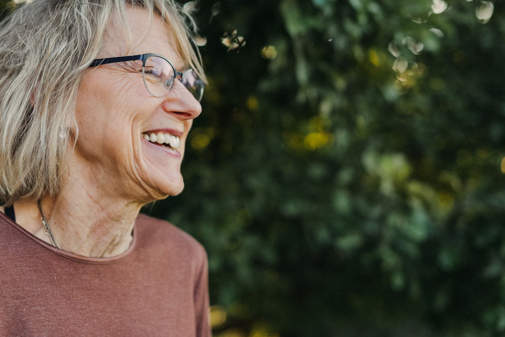 An older woman wearing glasses is smiling in front of a tree.