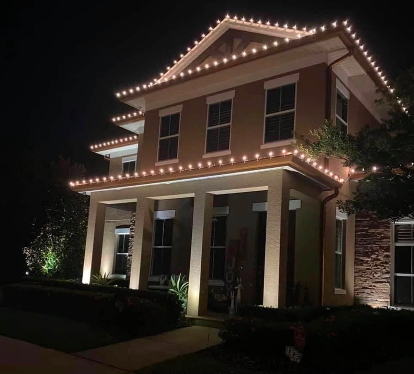 Two-story house at night outlined with warm white roof lights and lit columns in a dark yard