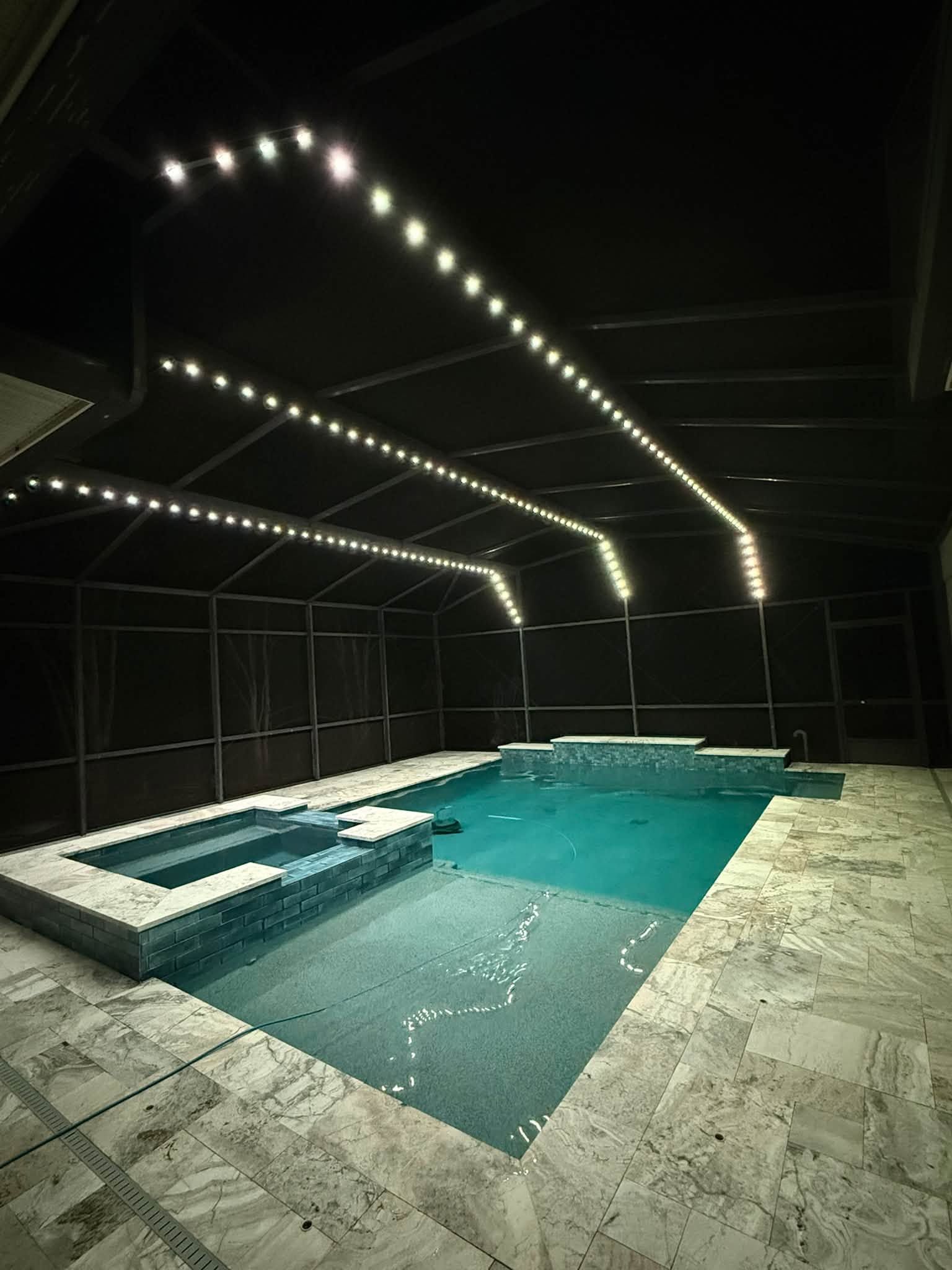 Modern indoor pool with turquoise water, white stone deck, and geometric overhead string lights at night