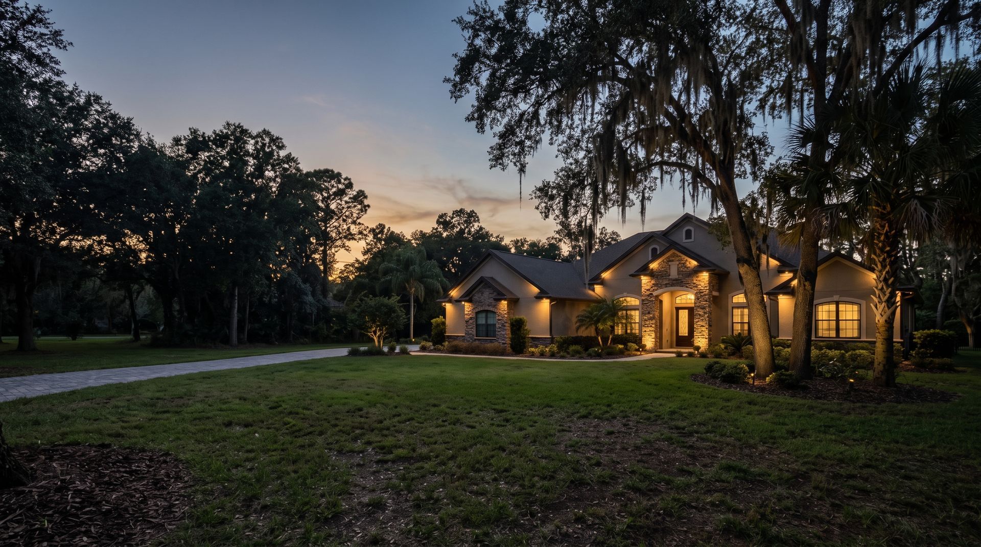 Suburban house at dusk with warm porch lights, surrounded by trees and a grassy yard