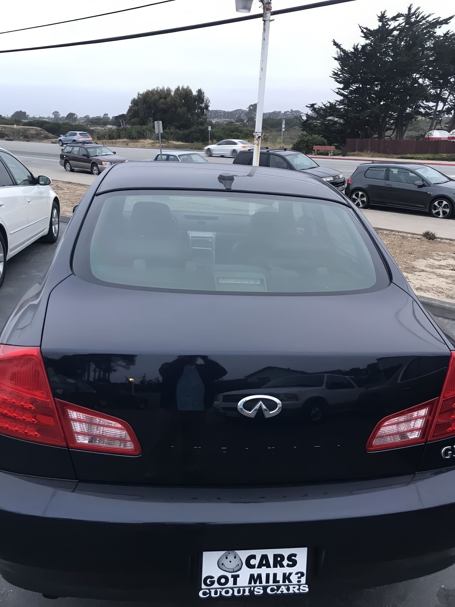 Rear view of a black Infiniti sedan in a parking lot, with cars and a road in the background