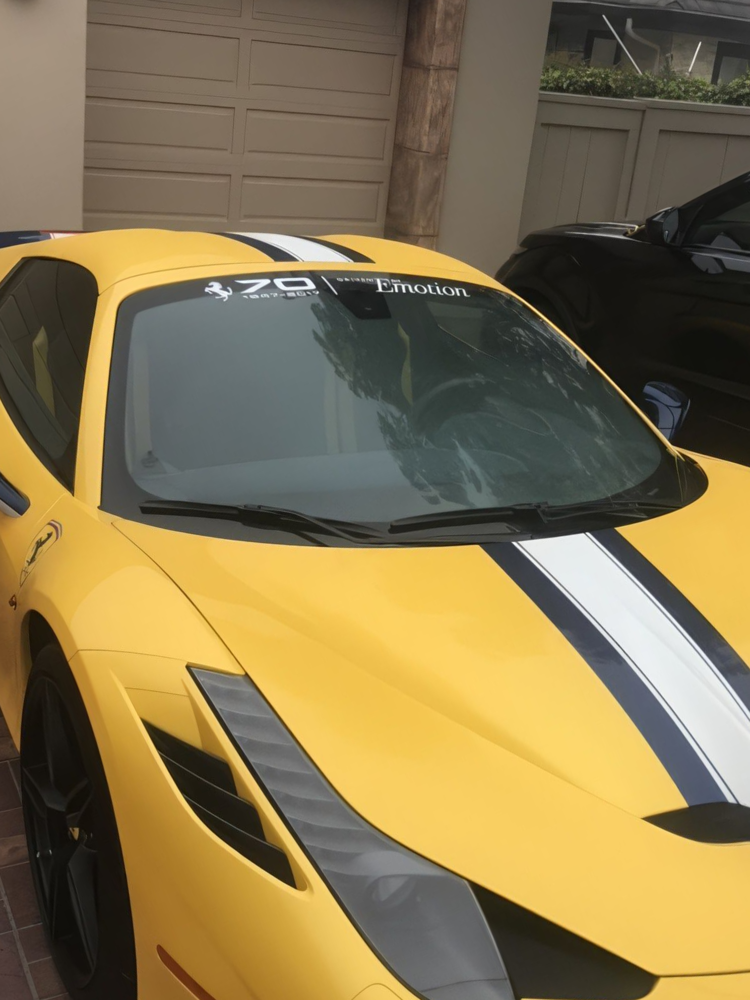 Yellow sports car with black racing stripes parked in a driveway near a garage