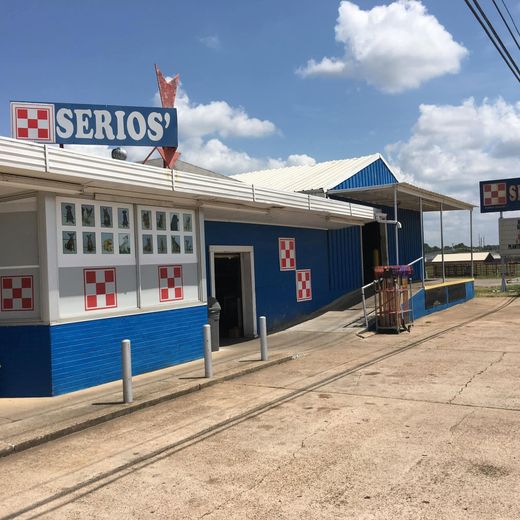 Small blue-and-white storefront with a SERIOS sign, tiled windows, and a dirt road under a sunny sky