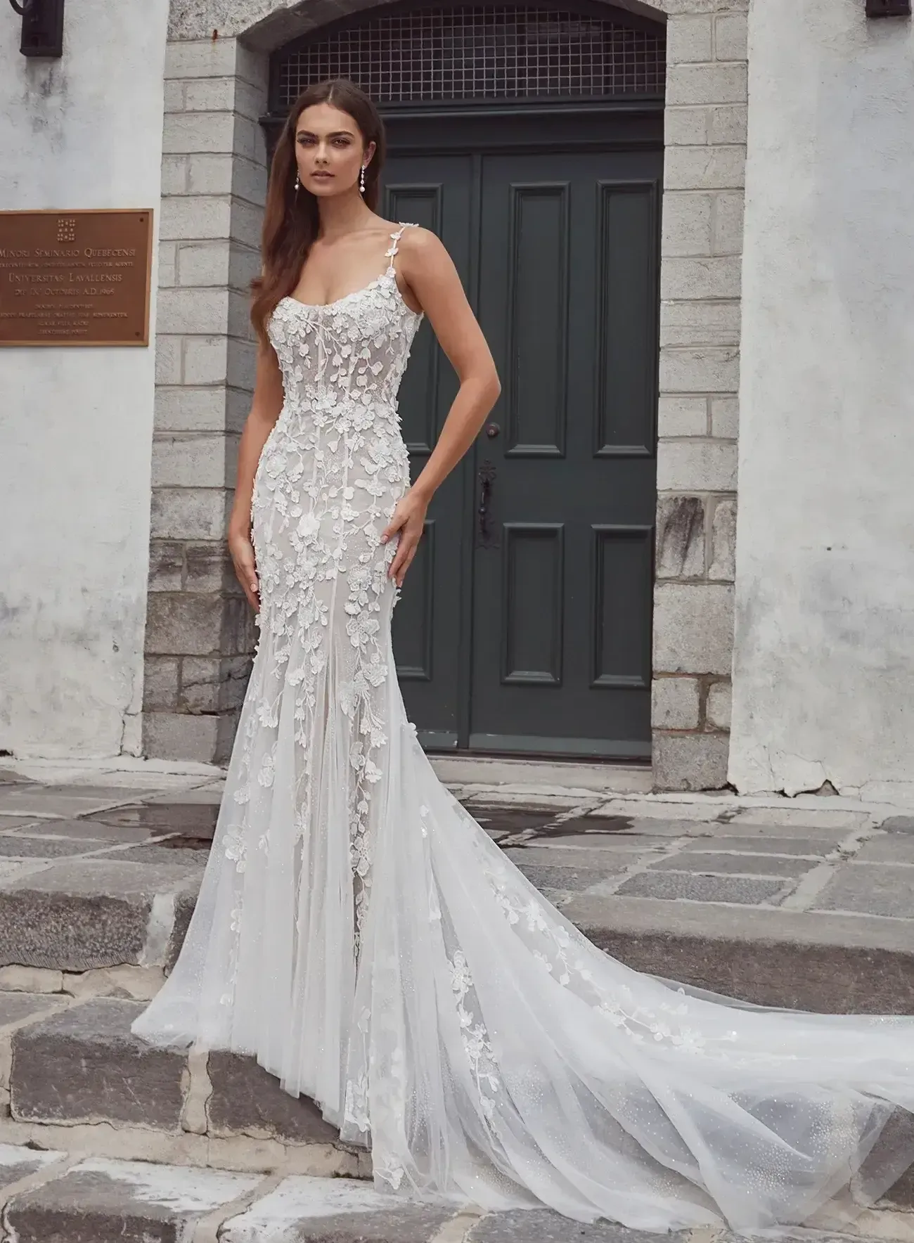 Woman in a white floral wedding dress stands on stone steps, train flowing.