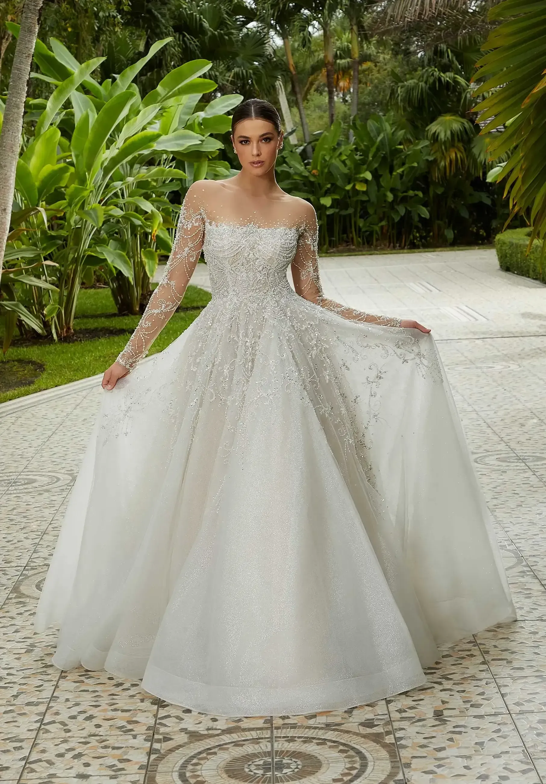 Woman in a sparkly white ballgown with long sleeves, standing on a mosaic path, in a garden.
