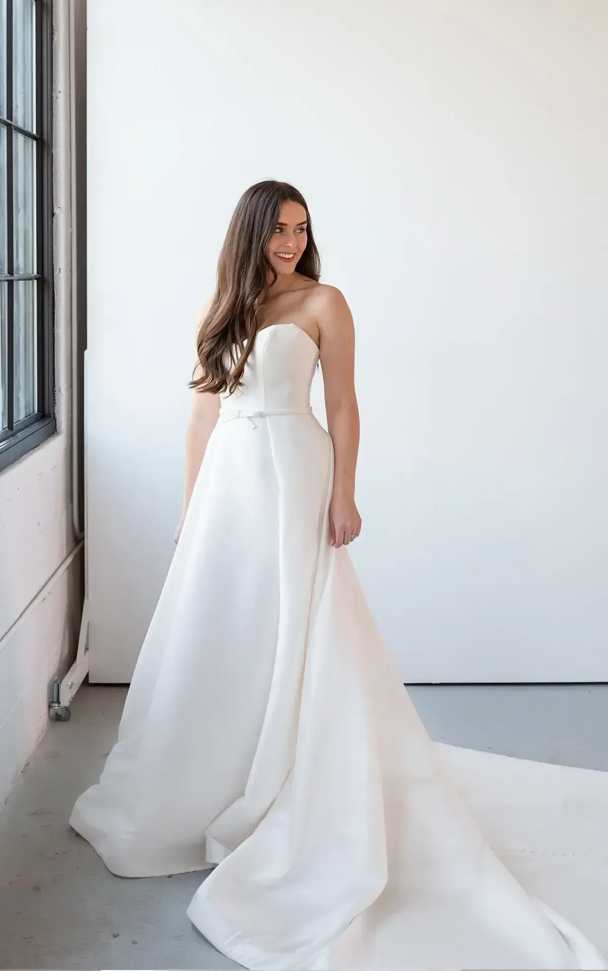 Woman in strapless white wedding dress with long train, smiling. Standing near a window, white wall backdrop.