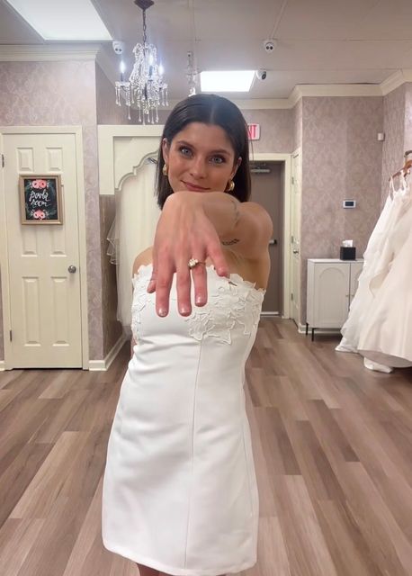 Woman in a white strapless dress reaching a hand toward the camera in a bridal salon