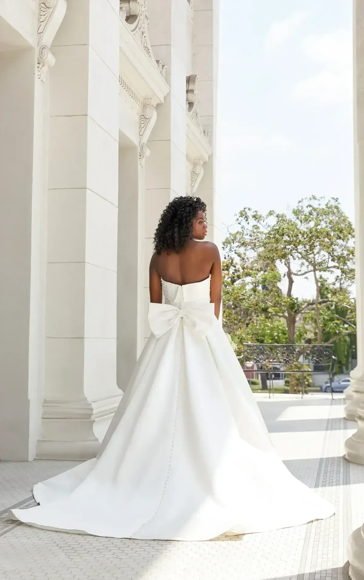 Woman in a white strapless wedding dress with a large bow, standing by columns.