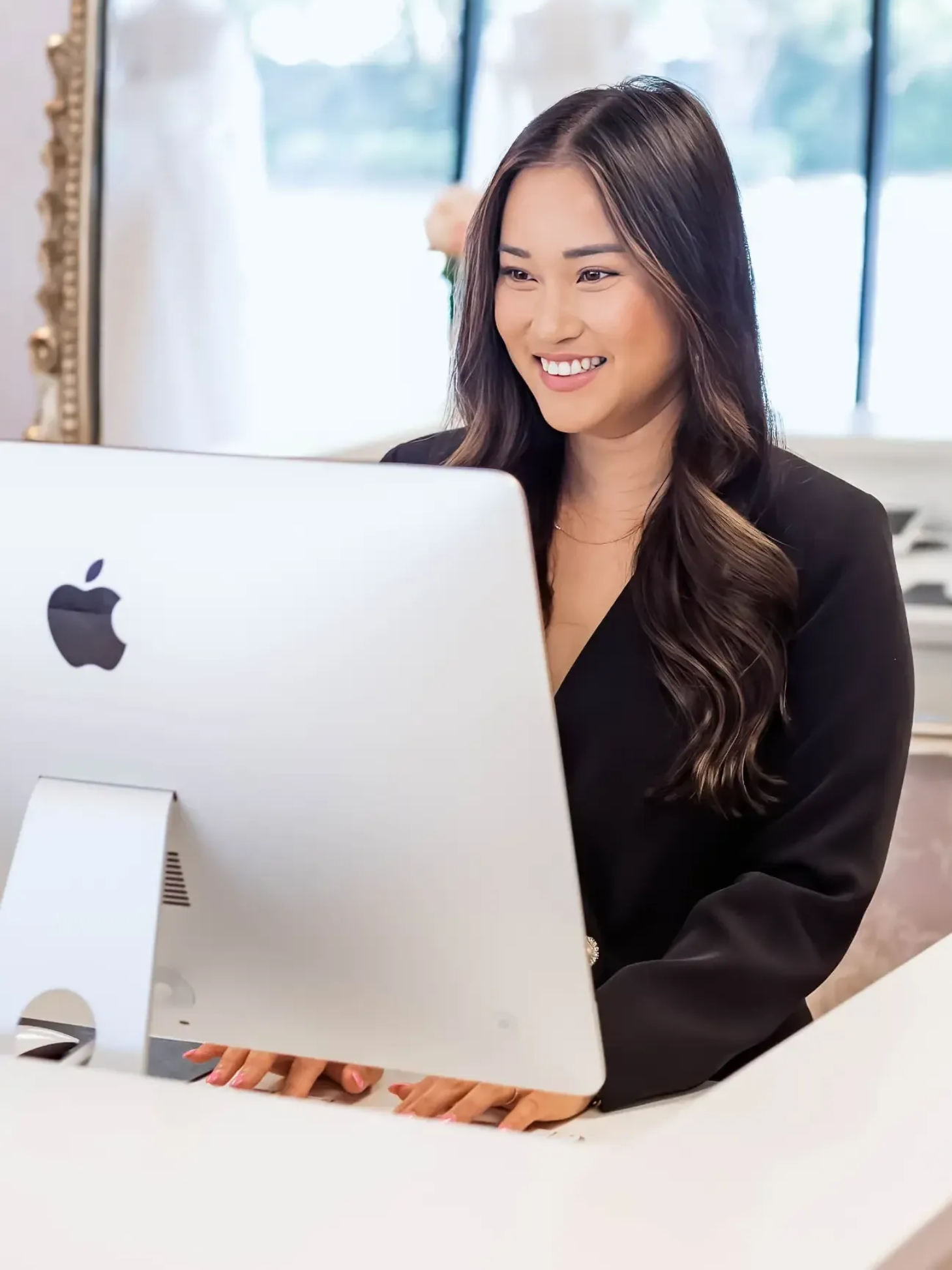 Woman smiling, looking at computer. Wearing a black blazer, white desk, wedding dress in background.