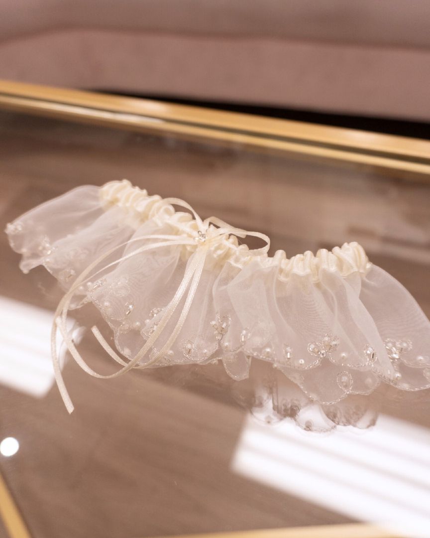 Ivory lace wedding garter with delicate bow, lying on a reflective surface.