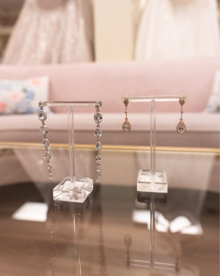 Two pairs of earrings on clear stands, reflecting on a glass surface. Soft pink couch in background.