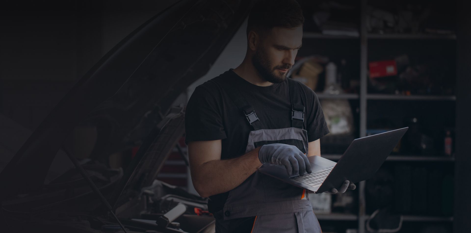 Mechanic inspecting a laptop in a dark garage beside a car with the hood open | PC Auto Repair