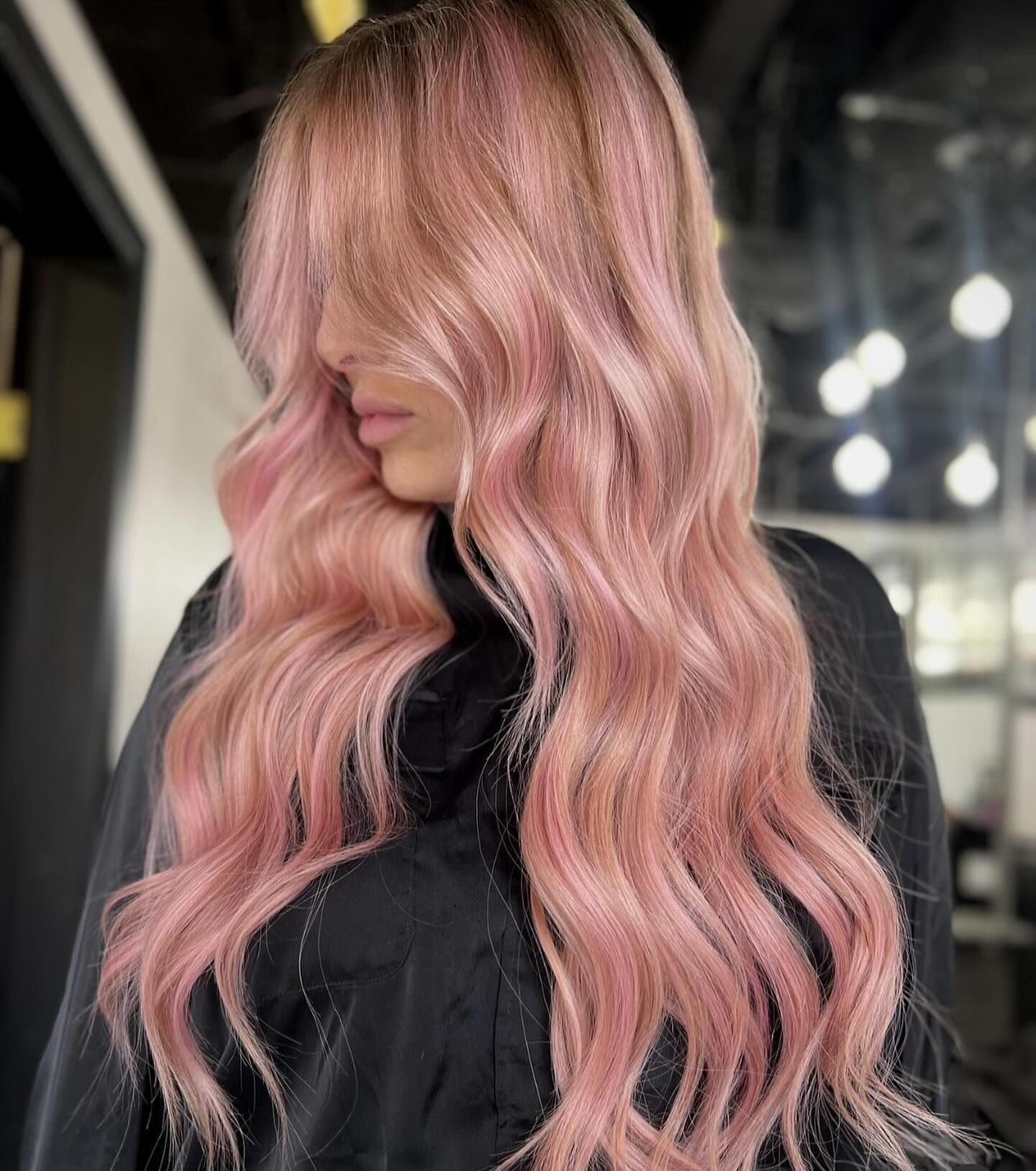 A person with long, wavy, pastel pink hair styled with loose curls, wearing a dark top in an indoor salon setting.