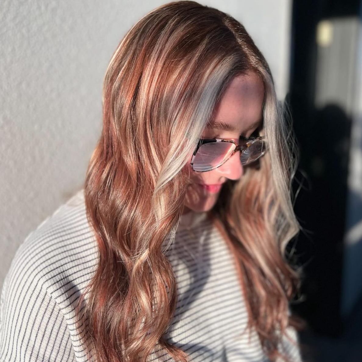 A person with wavy, auburn-toned hair featuring bright blonde face-framing highlights, wearing glasses and a striped top.