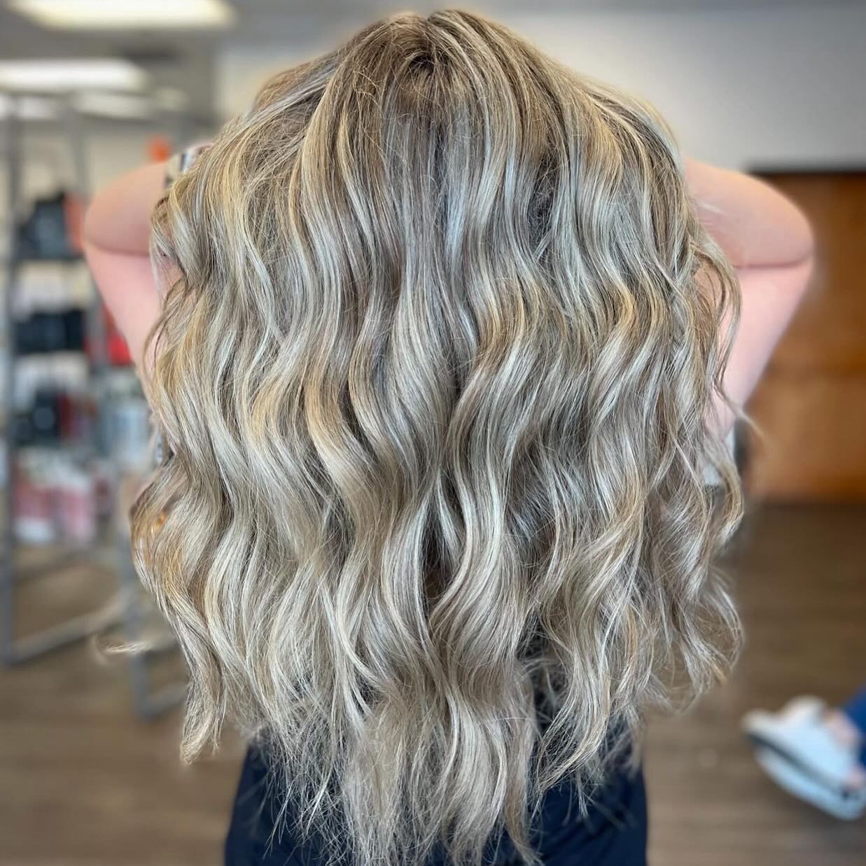 A person from behind displaying long, wavy blonde hair with subtle highlights, styled in a salon setting.