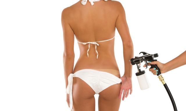 A person in a white bikini receiving a professional spray tan application from a handheld spray gun.