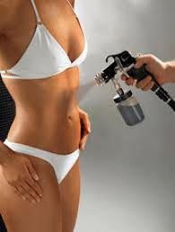 A hand uses a professional spray tanning machine to apply a fine mist onto a person's torso.