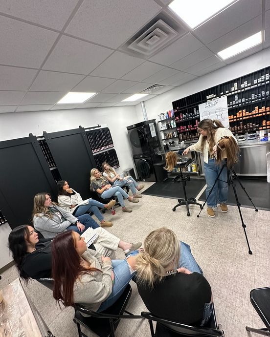 An instructor demonstrates a haircut on a mannequin head before a class of students sitting in a training room.