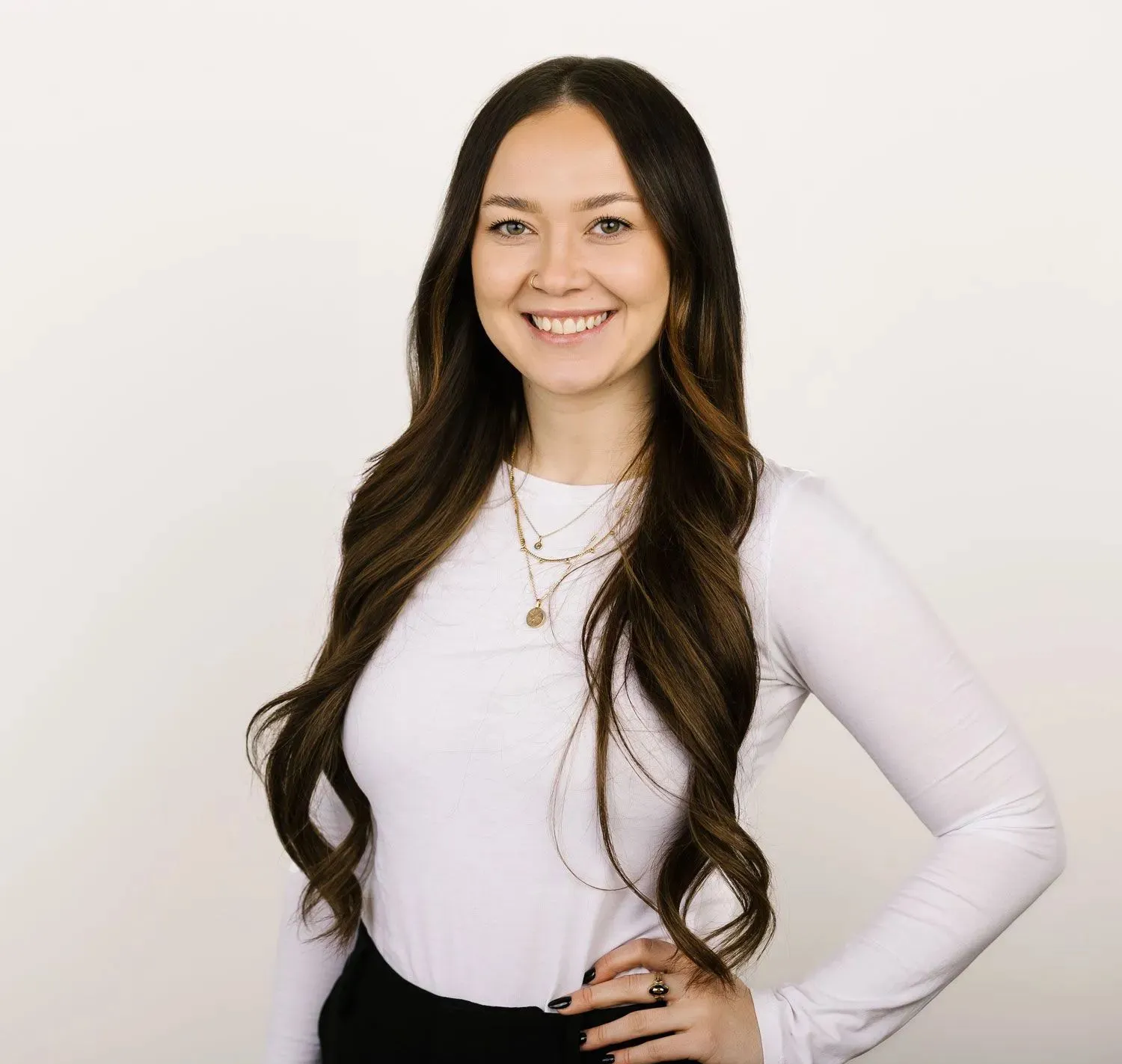 A person with long, dark wavy hair wearing a long-sleeved white top and layered gold necklaces, smiling against a white wall.