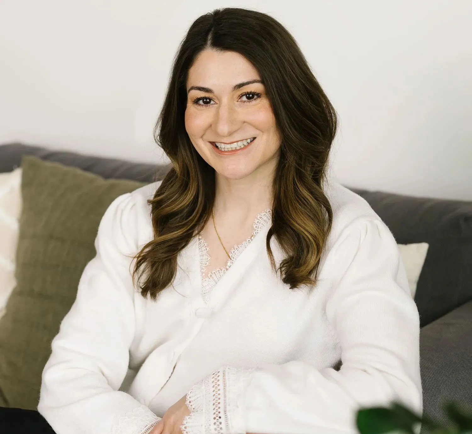A smiling person with long brown hair wearing a white lace-trimmed top, sitting on a sofa with olive and white pillows.