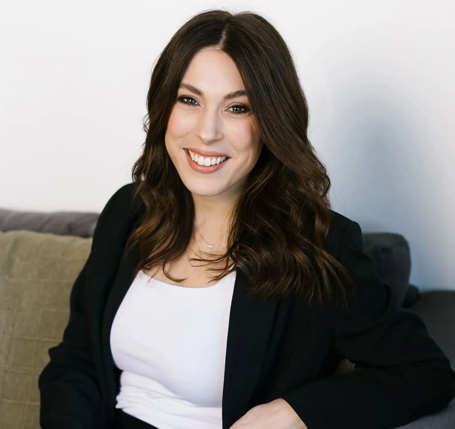 A woman with long brown hair, wearing a white shirt and black blazer, smiling while sitting on a couch.