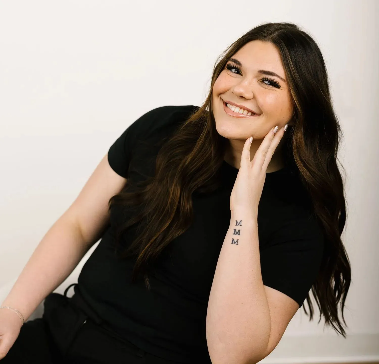 Smiling person with long brown hair, wearing a black shirt, posing against a white background with a hand on their face.