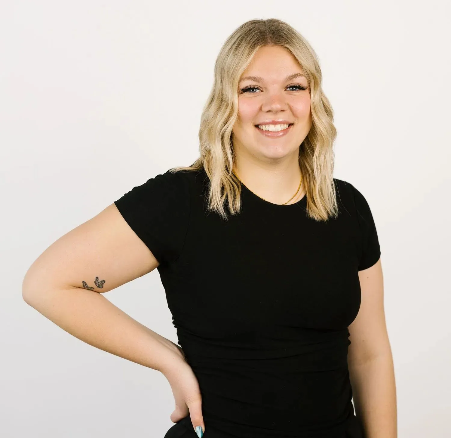 A person with shoulder-length blonde wavy hair wearing a black t-shirt, smiling against a plain white background.